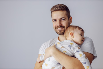 Young smiling attractive unshaven father finally succeeded to put his beloved son to sleep. Father is holding toddler and looking at camera.