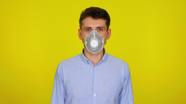 Young man in a protective mask looks at camera on yellow background with copy space. Epidemic precautions. Acute Respiratory Virus Infection. Illness cold flu. 4K footage.