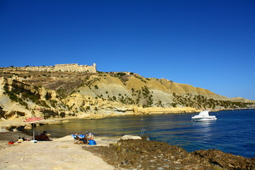 A rocky beach in Gozo, Malta, with sunbathers under the crystal clear blue sky.