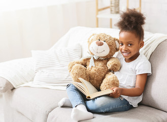 Cheerful black baby girl reading book on couch at home