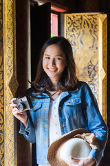 Close up portrait smiling young Asian woman in white dress holding and straw hat holding camera next Thai style pattern window