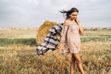 Full length portrait of a beautiful brunette in a dress and with a warm plaid. Woman enjoying a walk in a wheat field with hay bales