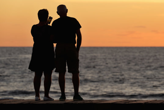 Silhouette of an older couple on the ocean coast as they take pictures of the sunset