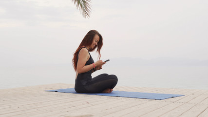 Fototapeta na wymiar Handsome female using her smartphone sitting on yoga mat at sea on foggy sunrise