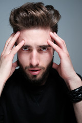 A young dark-haired man with a beard holds his head and shows different human emotions: hatred, fear, despair, horror, malaise, headache, clairvoyance. Close-up studio portrait of a man.