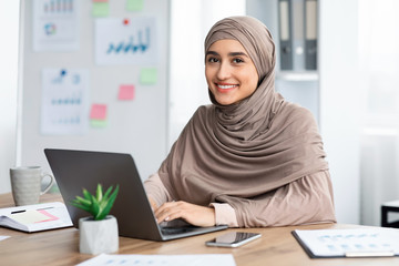 Fototapeta na wymiar Pretty arabic woman sitting at desk in office and using laptop