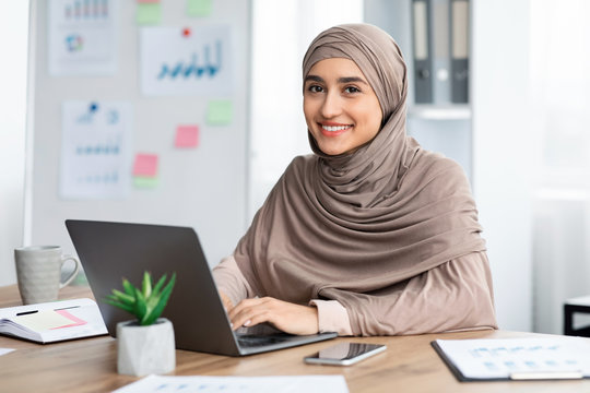Pretty arabic woman sitting at desk in office and using laptop - Powered by Adobe