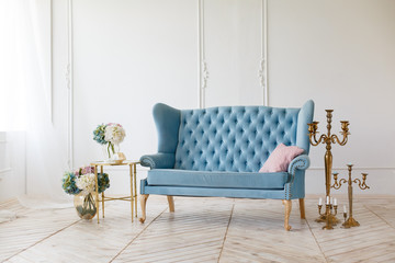 White living room with elegant blue classic sofa with golden candles and flowers. Royal light room. Beautiful armchair. Classic elegant interior with blue and white colors. Guest room, window light