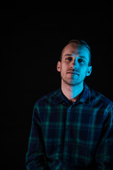 A man in a green checkered shirt looks to the camera on a black background. One half of the face is blue. Contrast lighting