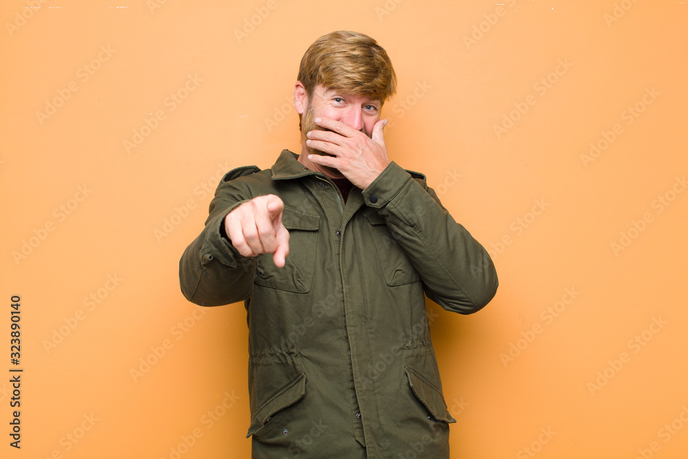 Wall mural young blonde man laughing at you, pointing to camera and making fun of or mocking you against flat wall