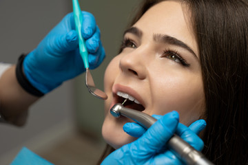dentist wearing blue gloves examines smiling woman patient's oral cavity using dental scaler and...