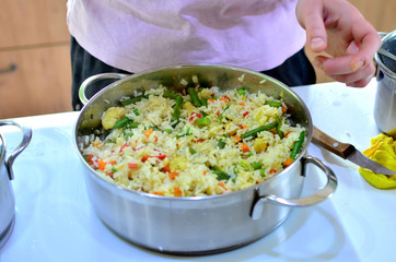 Woman cooks boiled rice with vegetables in a saucepan. Green beans, peppers, corn, peas, onions and carrots are prepared without loss of vitamins.
