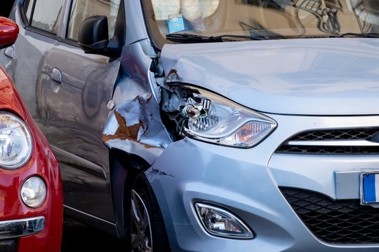 A small blue hatchback damaged in a car accident with a broken headlamp and fender waiting for repair