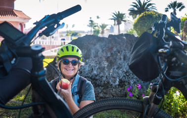 One elderly woman cyclist with yellow helmet have a break in the park . Sitting and eating a red apple. Moment of relaxation for a senior people