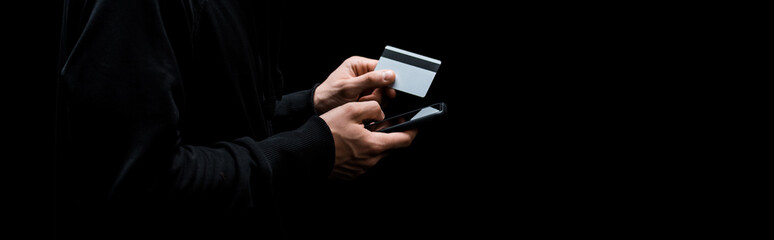 panoramic shot of hacker using smartphone and holding credit card isolated on black