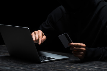cropped view of hacker using laptop while holding credit card isolated on black
