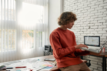Professional web illustrator. Side view of a young man in casual wear working with laptop while leaning on a table in the modern office. Creative people