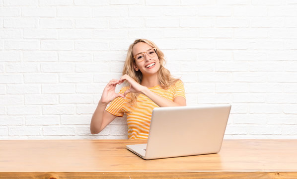 Young Blonde Woman Smiling And Feeling Happy, Cute, Romantic And In Love, Making Heart Shape With Both Hands Using A Laptop