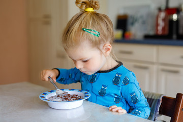 Gorgeous little toddler girl eating healthy cereal with milk for breakfast. Cute happy baby child in colorful clothes sitting in kitchen and having fun with preparing oats, cereals. Indoors at home