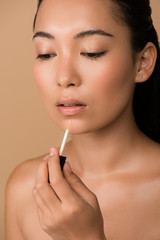 beautiful naked asian girl appying lip gloss on lips isolated on beige