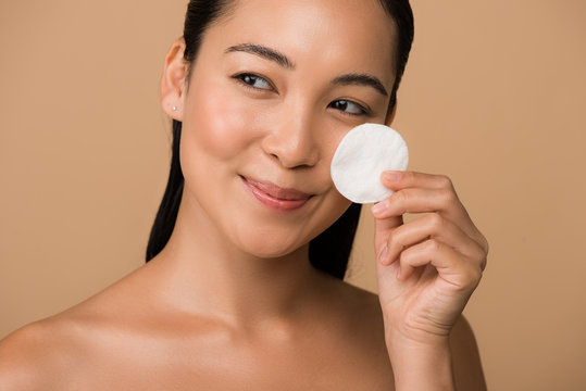 smiling beautiful naked asian girl holding cotton pad on face isolated on beige