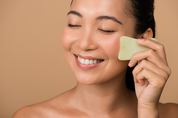 smiling beautiful naked asian girl with closed eyes using facial gua sha jade board isolated on...
