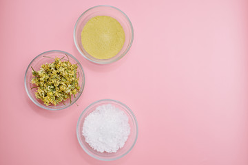 Obraz na płótnie Canvas Dried chamomile, yellow clay and sea salt in a glass bowl on a pink pastel background with copyspace. The concept of creating natural cosmetics and home personal care