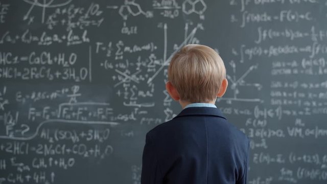 Back view of little boy in suit walking to chalkboard wall with formulas thinking looking at scientific calculations. Childhood, education and knowledge concept.