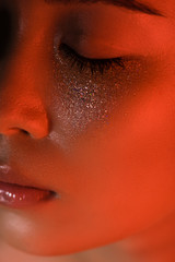 close up view of beautiful asian girl with silver sparkles on face and closed eyes in red lighting