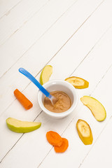 High angle view of baby food in bowl with spoon and sliced fruits and carrot on white wooden background