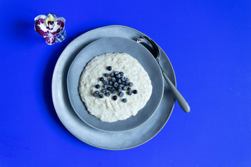 Oat meal with blackberry in gray plate with spoon on blue background, orchid flower