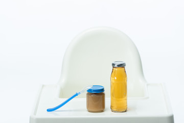 Jar of baby food with spoon and bottle of apple juice on table of feeding chair isolated on white