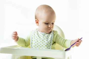 Cute baby holding spoon while sitting on feeding chair on white background
