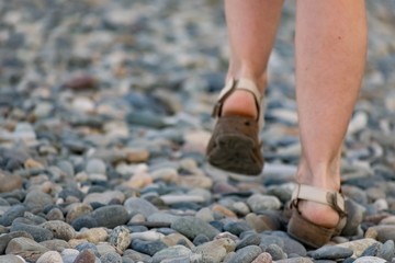 Women's feet in sandals walk along a stone road covered with smooth sea pebbles. White human skin and brown shoes like sandals
