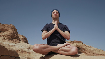 Fototapeta na wymiar Attractive male sitting in lotus pose and doing namaste on a rock in desert