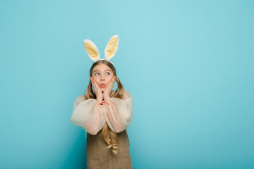 surprised kid with bunny ears touching face and looking away isolated on blue