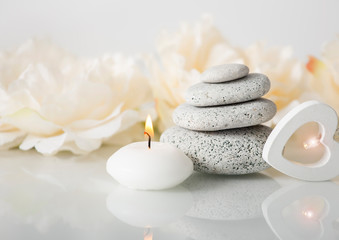 Fototapeta na wymiar Stacked zen stones with white burning candle and blurred soft floral background, white glowing wooden heart light. Relaxing lifestyle concept.