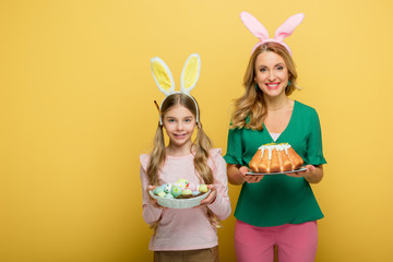 happy mother with bunny ears holding easter cake near daughter with painted chicken eggs isolated on yellow