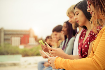 Teenagers texting mobile phone messages sitting on urban wall - Group of friends millennial using cellular outdoors - Concept of social network and telephone technology. Image
