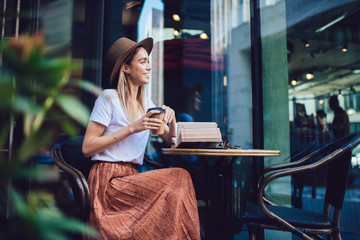 Joyful trendy woman drinking coffee while resting with book in cafe