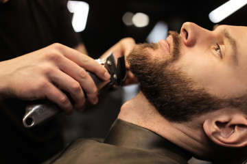 Hipster’s heaven. Close up photo of a barber using an electric shaver and a comb while trimming his customer’s beard.