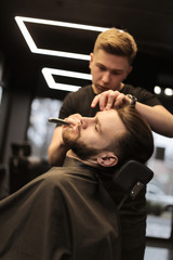 Looking up. Close-up photo of a professional hairstylist, who is grooming beard of his young client, who is looking in the mirror during the procedure.