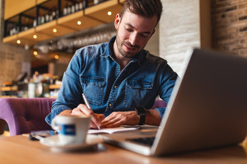 Obraz premium Freelancer working at cafe sitting at the table using laptop and writes in a notebook.
