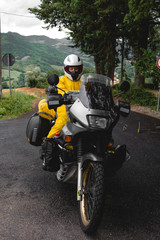 Motorcyclist traveler girl wearing yellow raincoat and sitting in a motorcycle saddle. Motorbike on mountain road. Extreme travel tour. Biker equipment. Vertical photo