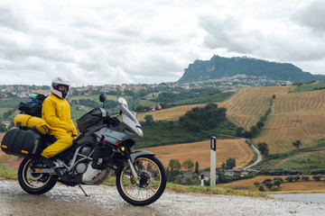 Fototapeta na wymiar Motorcyclist traveler girl wearing yellow raincoat and sitting in a motorcycle saddle. Motorbike on mountain road. Extreme travel tour. Biker equipment. View of the mountain. San Marino, Italy.