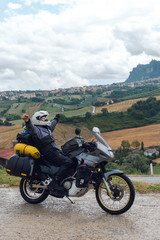 A motorcycle traveler arrived at his destination and he feels happiness. San Marino mountain on background, Adventure vacation, biker dressed in raincoat. Italy, Europe road trip. Vertical photo