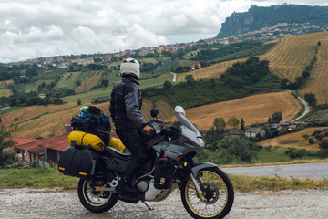 A motorcycle driver arrived at his destination and look distance San Marino mountain on background, Adventure vacation, biker dressed in raincoat. Italy, Europe road trip