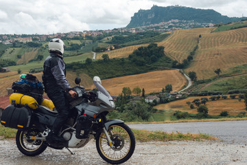 A motorcycle driver arrived at his destination and look distance San Marino mountain on background, Adventure vacation, biker dressed in raincoat. Italy, Europe road trip, copy space