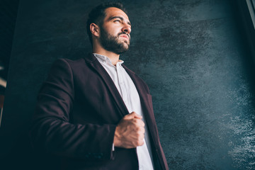 Serious caucasian prosperous male owner dressed in trendy expensive outfit looking away thoughtfully, handsome successful confident businessman posing near wall promotional background indoors.
