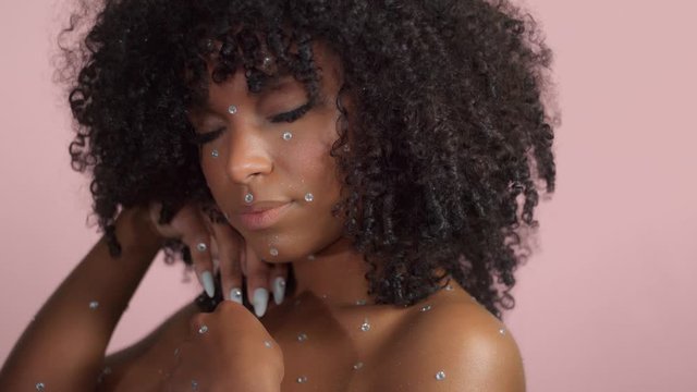 Mixed race black woman with curly hair covered by crystal makeup on pink background in studio touches her face and watching tot the camera